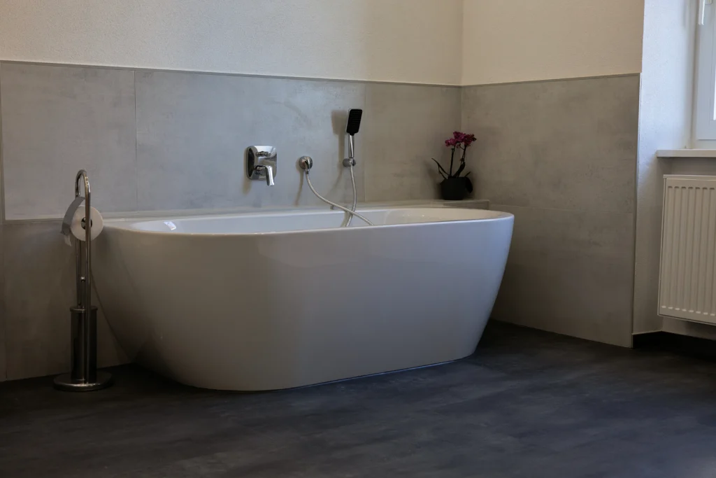 Freistehende Badewanne vor Wandfliesen in Betonoptik und dunklem Bodenbelag